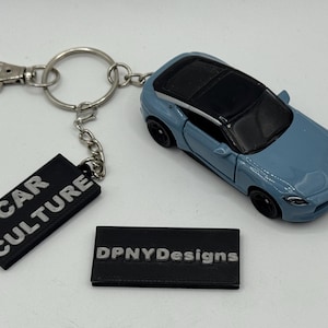 May include: A light blue diecast car keychain with a black roof. The keychain includes a black tag with "CAR CULTURE" in white lettering and a second black tag with "DPNYDesigns" in silver lettering.