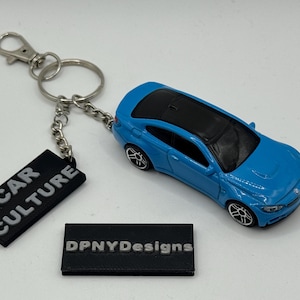 Puede incluir: Un llavero de coche de juguete azul con techo negro y detalles plateados. El llavero incluye una etiqueta negra que dice "CAR CULTURE". Una segunda etiqueta negra dice "DPNYDesigns".