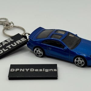 May include: A blue die-cast model car keychain with the text "CAR CULTURE" on a black rectangular tag. The car is a sports car with black wheels. A second black tag reads "DPNYDesigns".
