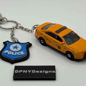 以下が含まれることがあります： 黒い車輪と黒いルーフラックを備えたオレンジ色の玩具タクシーが、警察のバッジキーホルダーの隣にあります。キーホルダーには、星が付いた青と白の警察バッジが付いています。「DPNYDesigns」というテキストが入った黒い長方形のオブジェクトも存在します。