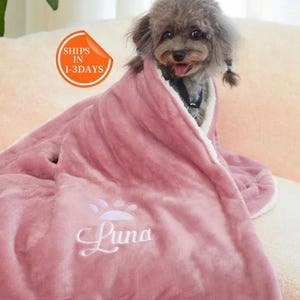 May include: A pink dog blanket with a white paw print and the name "Luna" embroidered on it. A small dog is curled up inside the blanket.