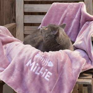 K&ouml;nnte beinhalten: Eine graue Katze liegt auf einer rosa Fleece-Decke mit dem Namen "Millie", der darauf gestickt ist. Die Decke hat einen wei&szlig;en Rand und ein Fischgr&auml;tenmuster.