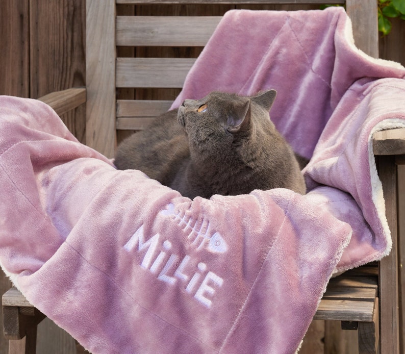 K&ouml;nnte beinhalten: Eine graue Katze liegt auf einer rosa Fleece-Decke mit dem Namen "Millie", der darauf gestickt ist. Die Decke hat einen wei&szlig;en Rand und ein Fischgr&auml;tenmuster.