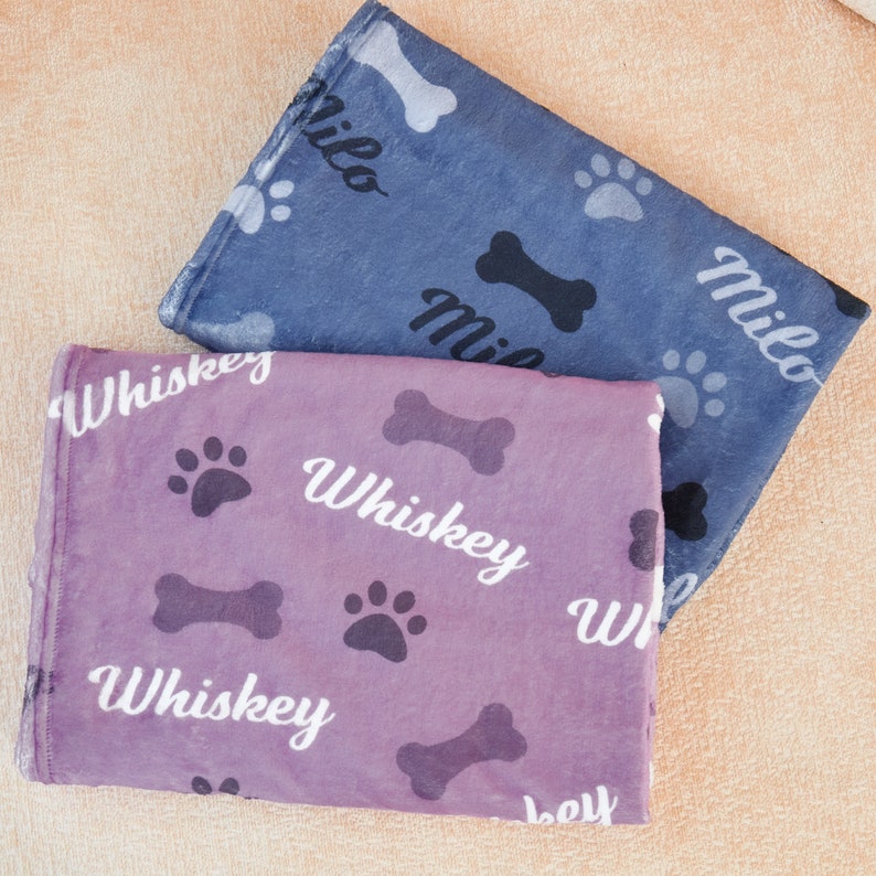 Puede incluir: Dos camas para perros con dise&ntilde;os de huellas de patas y huesos. Una cama es morada con el nombre "Whiskey" impreso en ella. La otra cama es azul con el nombre "Milo" impreso en ella.