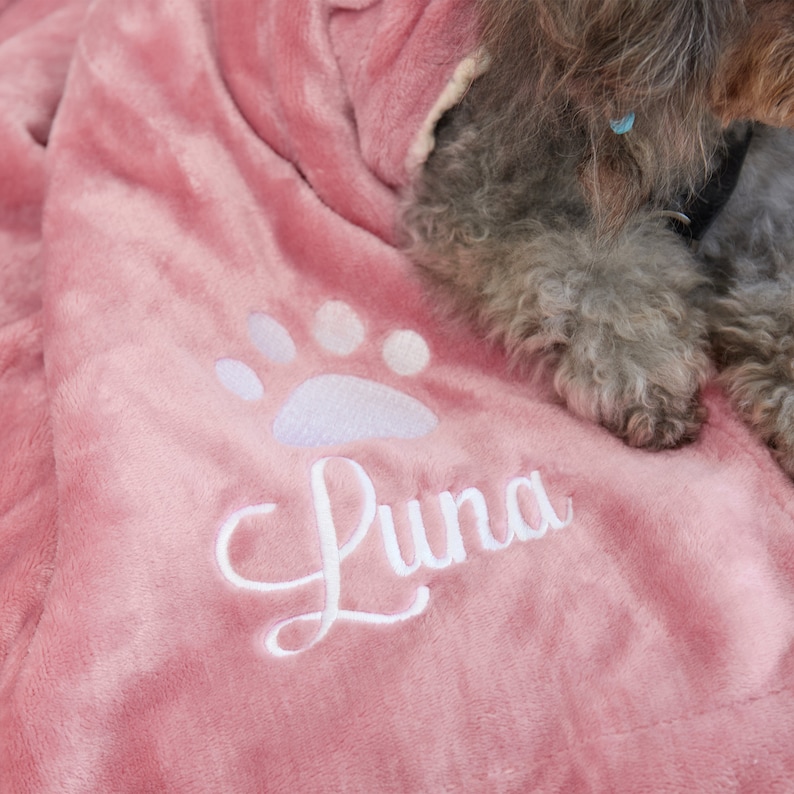 K&ouml;nnte beinhalten: Ein rosa Pl&uuml;sch-Hundebett mit einem wei&szlig;en Pfotenabdruck und dem Namen "Luna" bestickt.