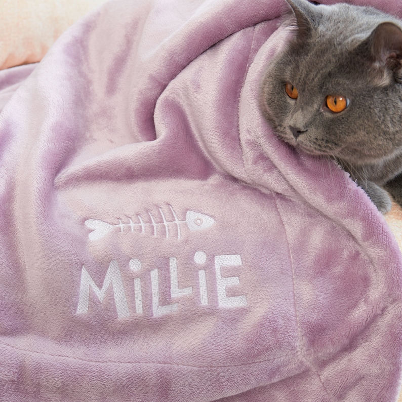 May include: A lavender-colored plush blanket with the name "Millie" embroidered in white, along with a fishbone design. A gray cat with orange eyes is partially visible, nestled in the blanket.