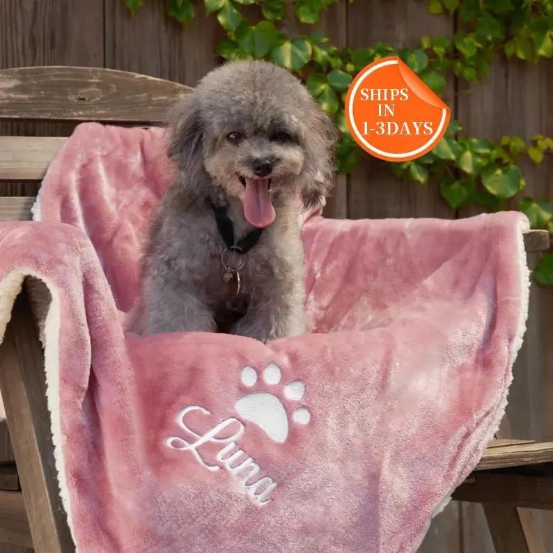 K&ouml;nnte beinhalten: Ein rosa-wei&szlig;es Pl&uuml;sch-Hundebett mit einem wei&szlig;en Pfotenabdruck und dem Namen "Luna" bestickt. Ein kleiner grauer Hund sitzt auf dem Bett.