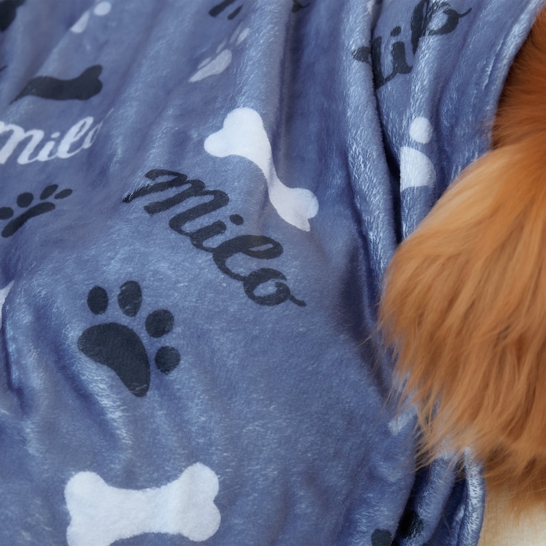 Puede incluir: Una manta de forro polar azul con un patr&oacute;n de huesos y huellas de patas blancas. La manta tiene el nombre "Milo" impreso en negro.