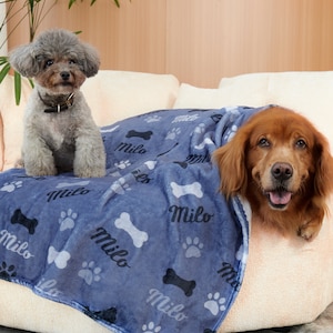 Puede incluir: Dos perros, un caniche gris y un golden retriever, se relajan en una manta de franela azul y gris con el nombre "Milo" impreso en ella. La manta tiene un dise&ntilde;o de huella de pata y hueso.