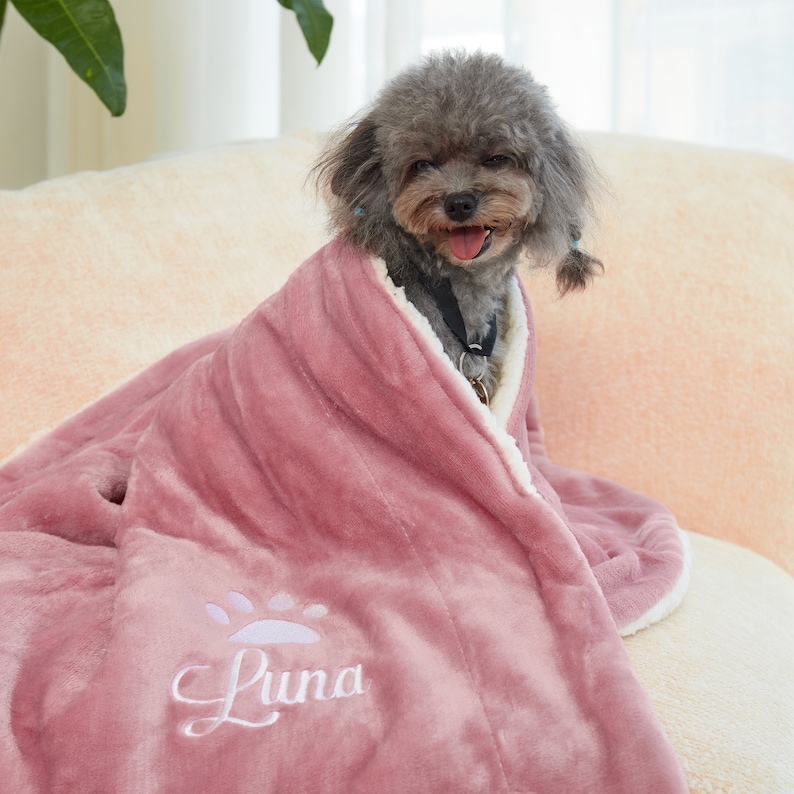 May include: A small, gray dog wrapped in a pink blanket with white trim. The blanket has a paw print design and the name "Luna" embroidered on it in white. The dog is sitting on a light-colored, plush surface.