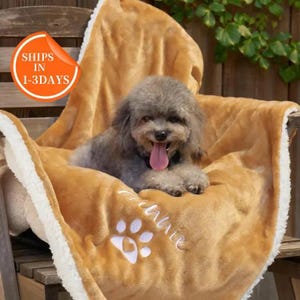 May include: A small, gray dog with its tongue out rests on a tan pet blanket. The blanket has a white paw print and the name "Sunshine" embroidered on it. The blanket is on a wooden chair.