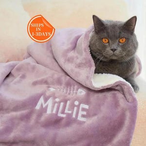 May include: A gray cat with orange eyes is wrapped in a lavender blanket. The blanket has a white fishbone design and the name "Millie" embroidered on it. The blanket is soft and plush.