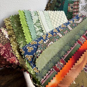 Puede incluir: Una colección de muestras de tela en varios tonos de verde, azul, naranja y marrón. Las piezas de tela tienen diseños florales y geométricos. Los bordes están cortados con un patrón en zigzag. Las muestras están dispuestas en forma de abanico.
