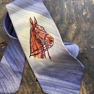 May include: A blue necktie featuring a diagonal light-colored band across the center. The tie displays a detailed illustration of a brown horse head wearing a red bridle, positioned centrally within the light-colored diagonal section.