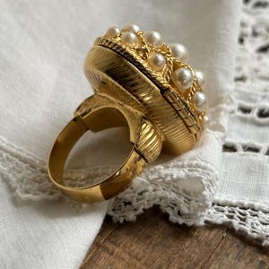 Puede incluir: Un anillo de tono dorado con un engaste circular grande y texturizado, coronado por un grupo de pequeñas perlas blancas redondas. La banda y los lados del engaste presentan un patrón lineal fino y grabado, con un borde dorado trenzado.