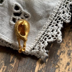 May include: A gold-toned metal brooch shaped like the rear view of a cat, featuring a textured finish on the fur and a curved tail. The brooch is pinned onto a piece of white fabric with intricate lace trim and decorative cut-out patterns.