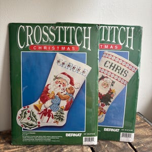 May include: Two sealed Bernat cross-stitch kits for Christmas stockings. The left kit features a Santa and friends design with the name Carole, measuring 9 inches by 15 inches. The right kit shows a Santa design with the name Chris.