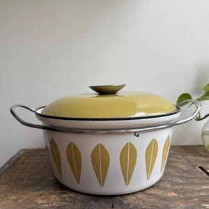 Puede incluir: Una olla de esmalte blanco vintage con tapa amarilla y estampado de hojas. La olla tiene asas plateadas y un borde negro. Una pequeña planta verde en un jarrón de cristal está en el fondo.