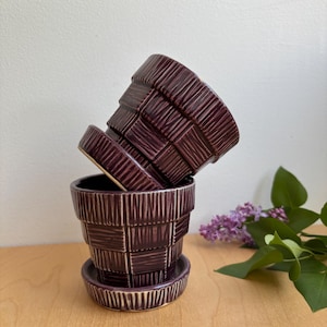 May include: Three plum-coloured ceramic flower pots with a textured, basket-weave design. The pots are stacked on a wooden surface, with a small saucer and a sprig of purple flowers in the background.