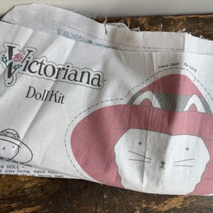 May include: A white fabric panel for a Victoriana Doll Kit featuring printed sewing patterns. The fabric displays a large cat face wearing a wide-brimmed red hat with a dotted pattern, along with text instructions and a smaller line drawing of the cat.