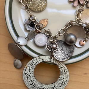 May include: A silver-toned statement necklace with various charms and pendants. The necklace features a large, openwork circular pendant, a variety of smaller charms, and a chunky chain. The charms include floral, round, and textured elements.