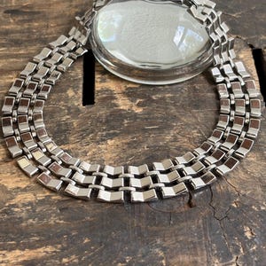 May include: A multi-strand necklace featuring three rows of polished silver-tone metal links. Each link has a rectangular shape with a slightly angled, stepped surface. The necklace is arranged in a circular formation, showcasing a consistent metallic finish and a structured, repeating pattern throughout the entire length of the piece.
