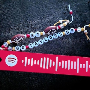 May include: A red keychain with a white Spotify barcode and the text "GO TAYLORS BF 87" in white beads with brown footballs and red, white, and yellow beads.