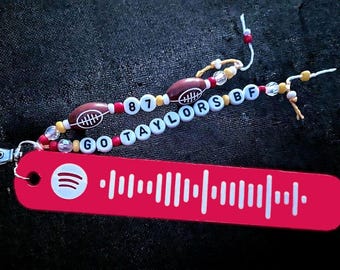 Pode incluir: Um chaveiro vermelho com um código de barras Spotify branco e o texto "GO TAYLORS BF 87" em contas brancas com bolas de futebol marrons e contas vermelhas, brancas e amarelas.