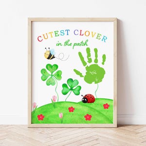 May include: A framed watercolor print featuring a whimsical garden scene. The artwork includes a green handprint, clovers, a ladybug, and a bee. The text "Cutest Clover in the patch" is written in rainbow colors. The print is set against a white background.