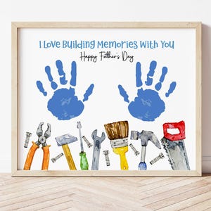 Könnte beinhalten: Gerahmtes Kunstwerk mit dem Text "I Love Building Memories With You Happy Father's Day". Der Druck zeigt zwei blaue Handabdrücke über Aquarellillustrationen von Werkzeugen, darunter eine Zange, ein Hammer und eine Säge. Der Rahmen ist hellbraun.