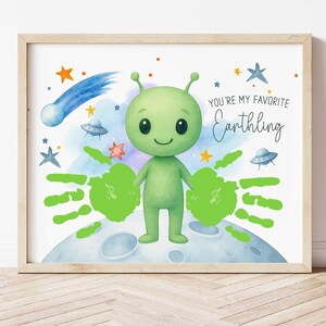 Pode incluir: Impressão emoldurada com um personagem alienígena verde e impressões de mãos em um planeta. A obra de arte inclui um cometa, estrelas e o texto "You're my favorite Earthling". A impressão está em um fundo branco.