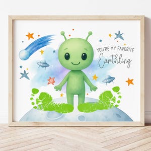 Pode incluir: Uma impressão emoldurada em aquarela com um alienígena verde sorridente em uma lua. A impressão inclui um cometa azul, estrelas e o texto "You're my favorite Earthling".