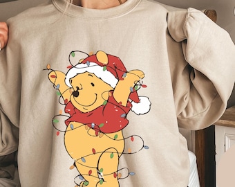 May include: A beige sweatshirt with a Winnie the Pooh graphic. Pooh is wearing a Santa hat and is surrounded by Christmas lights. The text "SAND" is visible in the lower right corner of the image.