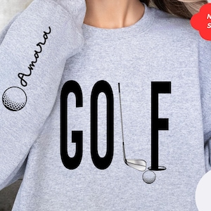 Pu&ograve; includere: Felpa grigia con la parola "GOLF" stampata in nero. La parola &egrave; stilizzata con una mazza da golf e una palla. La manica ha la parola "Amarra" stampata in corsivo.