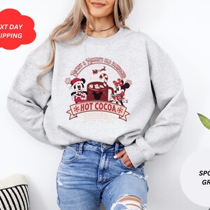 May include: A gray sweatshirt with a Christmas-themed design featuring Mickey Mouse and Minnie Mouse holding a cup of hot cocoa. The text on the sweatshirt reads "Mickey & Minnie Old Fashioned Hot Cocoa".