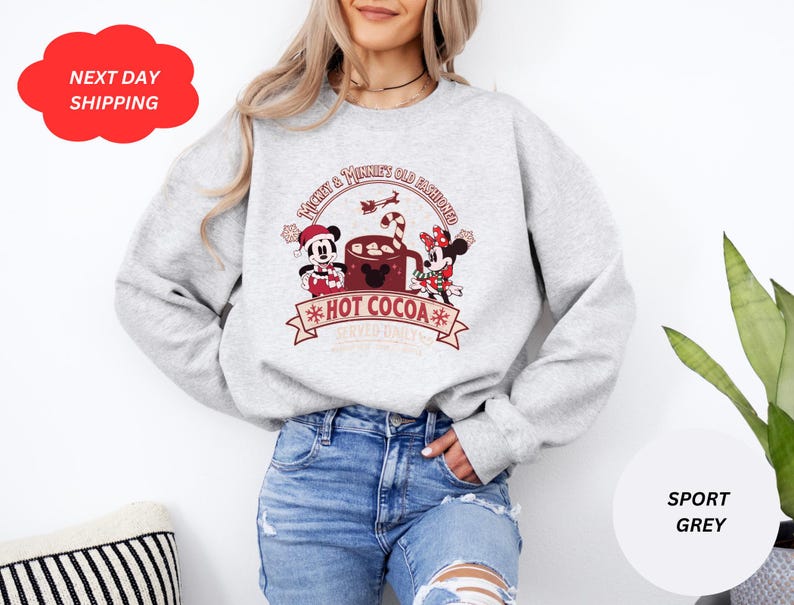 May include: A gray sweatshirt with a Christmas-themed design featuring Mickey Mouse and Minnie Mouse holding a cup of hot cocoa. The text on the sweatshirt reads "Mickey & Minnie Old Fashioned Hot Cocoa".