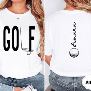 May include: White t-shirt with the word "GOLF" in large black letters on the front, along with a golf club, ball, and hole graphic. The back of the shirt features the name "Amara" in a vertical script above a golf ball graphic.