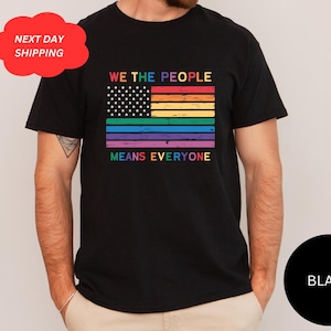 以下が含まれることがあります： 虹色の旗のデザインと「WE THE PEOPLE MEANS EVERYONE」の文字が入った黒いTシャツ。旗には虹色のストライプと星が含まれており、包括性を象徴しています。赤い雲型のグラフィックは「NEXT DAY SHIPPING」を示しています。