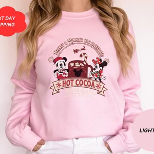 May include: Light pink sweatshirt with a graphic of Mickey Mouse and Minnie Mouse holding a mug of hot cocoa. The text on the sweatshirt reads "Mickey & Minnie's Old Fashioned *Hot Cocoa* Served Daily".