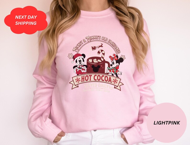 May include: Light pink sweatshirt with a graphic of Mickey Mouse and Minnie Mouse holding a mug of hot cocoa. The text on the sweatshirt reads "Mickey & Minnie's Old Fashioned *Hot Cocoa* Served Daily".