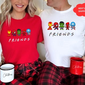 Puede incluir: Dos tazas de Navidad rojas y blancas con la palabra "Navidad" escrita en ellas. Una pareja lleva camisas rojas y blancas a juego con un gr&aacute;fico de superh&eacute;roes de dibujos animados y la palabra "FRIENDS" escrita debajo.