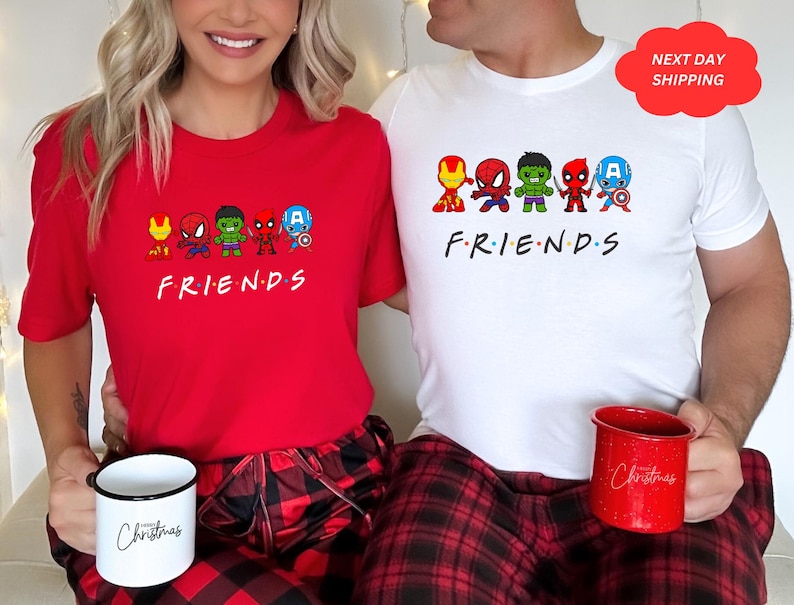 Puede incluir: Dos tazas de Navidad rojas y blancas con la palabra "Navidad" escrita en ellas. Una pareja lleva camisas rojas y blancas a juego con un gr&aacute;fico de superh&eacute;roes de dibujos animados y la palabra "FRIENDS" escrita debajo.