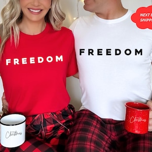 May include: A red t-shirt with the word "FREEDOM" in white, paired with red and black plaid pajama pants. A white mug with "Christmas" in black script is held. A white t-shirt with "FREEDOM" and a red mug with "Christmas" are also shown.
