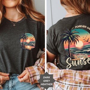 May include: Dark grey heather t-shirt featuring a colorful sunset and palm tree graphic. The front of the shirt has a small circular design, while the back displays a larger design with the words "Forever Chasing Sunsets."