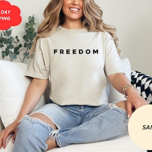 May include: Sand-colored t-shirt with the word "FREEDOM" printed in black. The model is wearing light blue ripped jeans. A red cloud-shaped sticker with the words "NEXT DAY SHIPPING" is visible in the upper left corner.