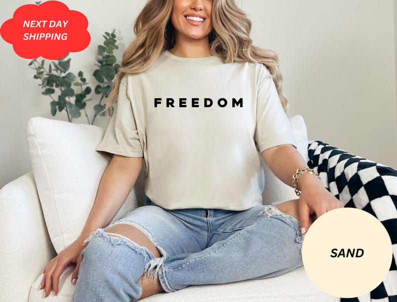 May include: Sand-colored t-shirt with the word "FREEDOM" printed in black. The model is wearing light blue ripped jeans. A red cloud-shaped sticker with the words "NEXT DAY SHIPPING" is visible in the upper left corner.