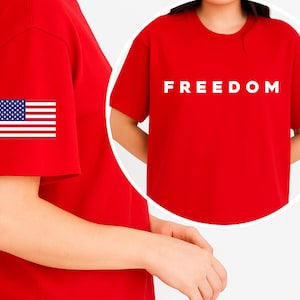 May include: A red t-shirt with the word "FREEDOM" printed in white across the chest. The left sleeve features a small American flag patch. The shirt is a solid red color.