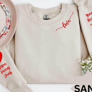 Puede incluir: Una sudadera de color arena con corazones rojos y la palabra "love" escrita en cursiva en la parte delantera. La manga izquierda tiene los nombres "Luna, Harry, Jack" escritos en rojo. La manga derecha tiene las palabras "you have my heart" escritas en rojo.