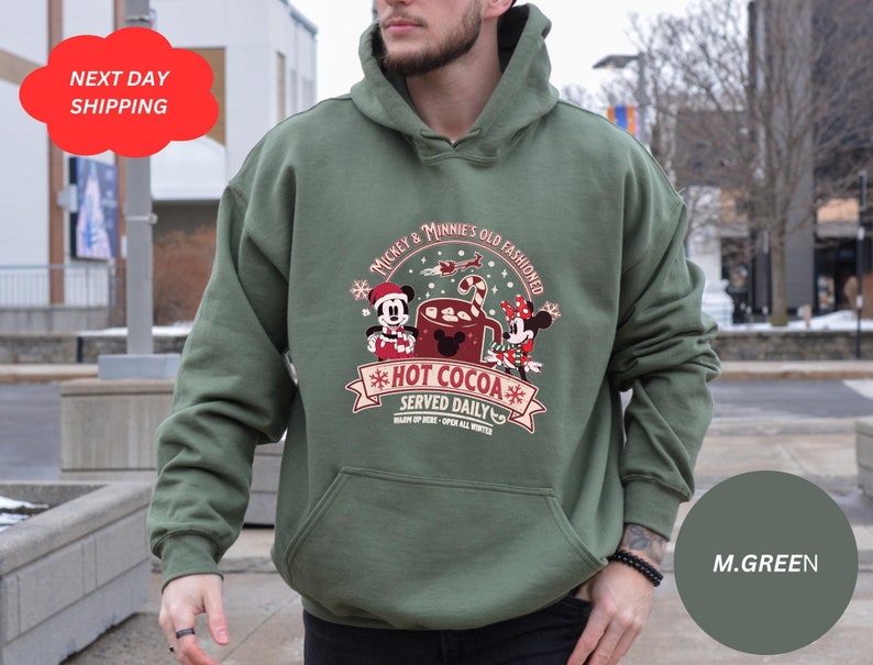 May include: Green hooded sweatshirt with a Christmas-themed design featuring Mickey and Minnie Mouse enjoying hot cocoa. The text reads "Mickey & Minnie's Old Fashioned *Hot Cocoa* Served Daily".