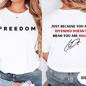 Op de afbeelding: Wit T-shirt met het woord "FREEDOM" in zwarte letters op de voorkant. De achterkant toont de tekst "JUST BECAUSE YOU ARE OFFENDED DOESN'T MEAN YOU ARE RIGHT" in zwart en rood, met een handtekening eronder.